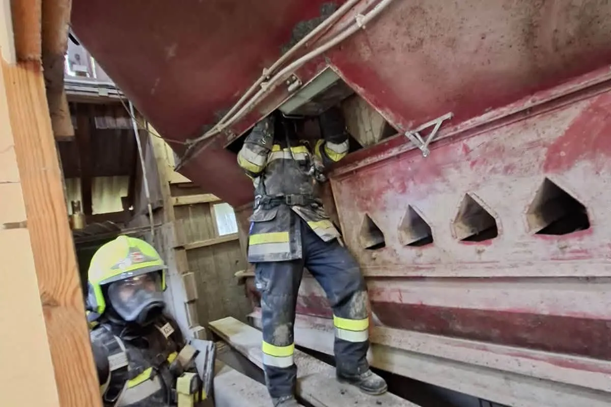 Zwei Feuerwehrleute in voller Ausrüstung arbeiten an einem großen roten Behälter. Das Bild zeigt eine industrielle Umgebung im Inneren eines Gebäudes.