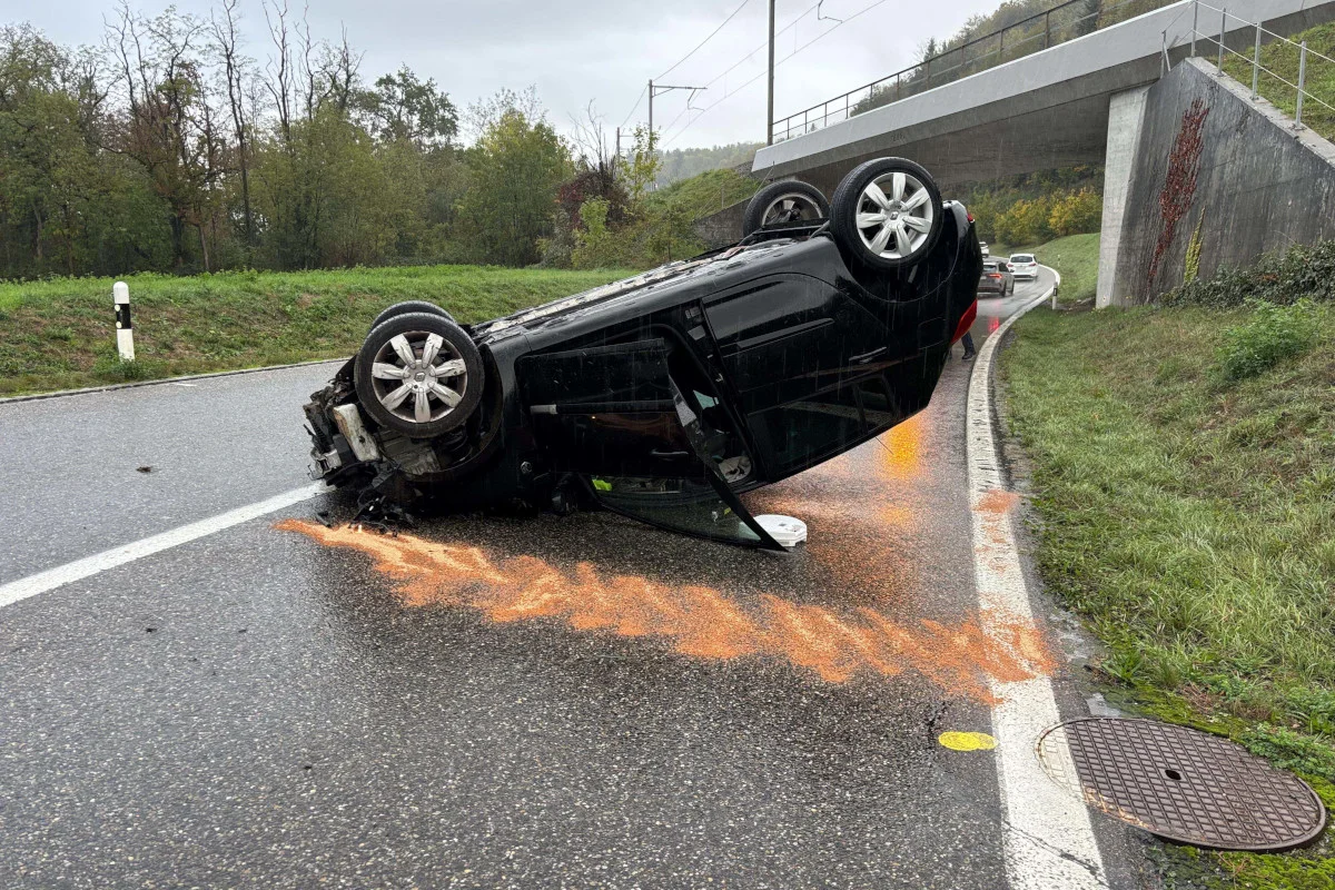 Ein Auto liegt auf dem Dach auf einer nassen Straße nahe einer Brücke. Im Vordergrund sieht man ausgelaufenes Öl und Glassplitter.