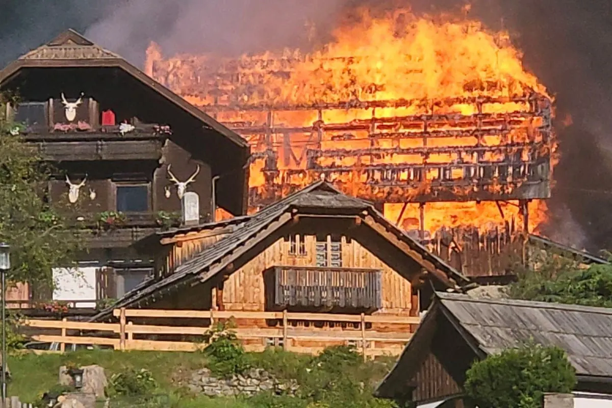Ein großer Gebäudebrand bei einem Bauernhof, die Flammen schlagen hoch und rauchen heftig. Vorne sind traditionelle Holzhäuser zu sehen.