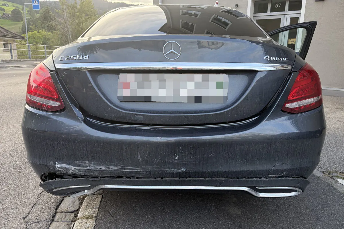 Ein dunkler Mercedes-Benz mit sichtbarem Schaden an der hinteren Stoßstange steht vor einem Gebäude auf der Straße.