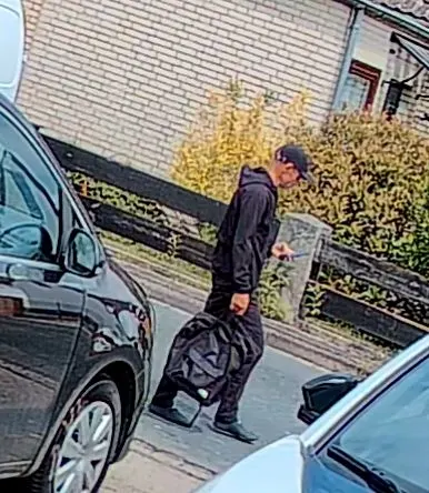 Ein Mann in dunkler Kleidung trägt eine Tasche und schaut auf sein Handy, während er an geparkten Autos vorbeigeht.