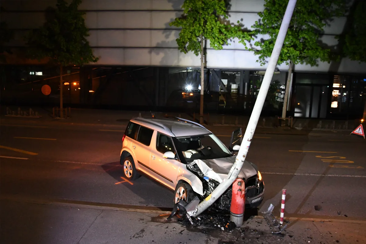 Ein silbernes Auto ist frontal gegen einen Laternenmast gefahren, der Mast ist stark geneigt. Eine nächtliche Straßenszene zeigt das beschädigte Fahrzeug und die Umgebung.