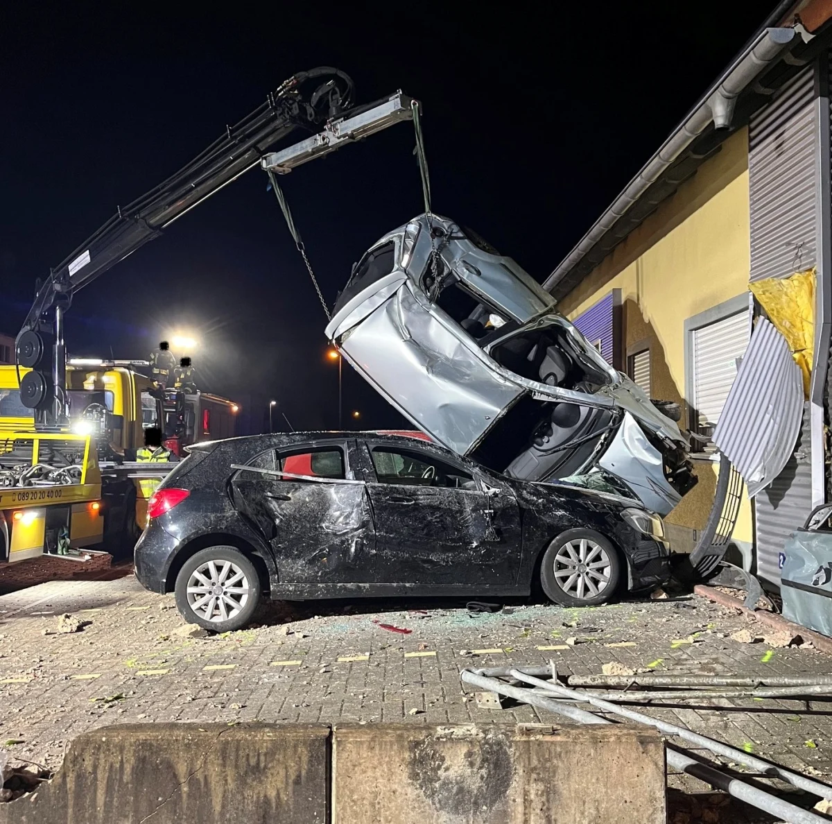 Zwei Fahrzeuge nach einem Unfall, wobei eines auf dem Dach des anderen gelandet ist. Ein Kran hebt das obere Auto an, um es zu bergen. Die Szene findet nachts statt.