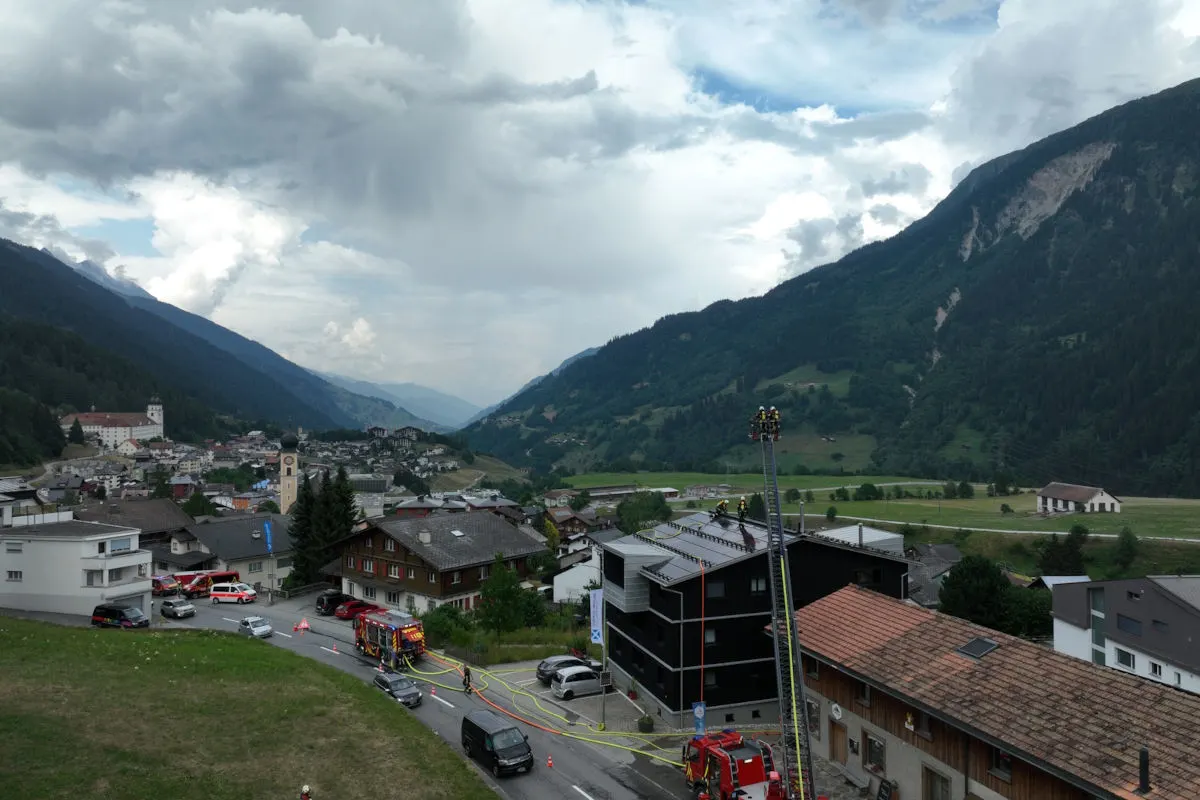 Mehrere Feuerwehrfahrzeuge und Einsatzkräfte sind bei einem Gebäude in einer alpinen Ortschaft im Einsatz. Eine Leiter ist auf das Dach ausgefahren, wo Feuerwehrleute arbeiten.