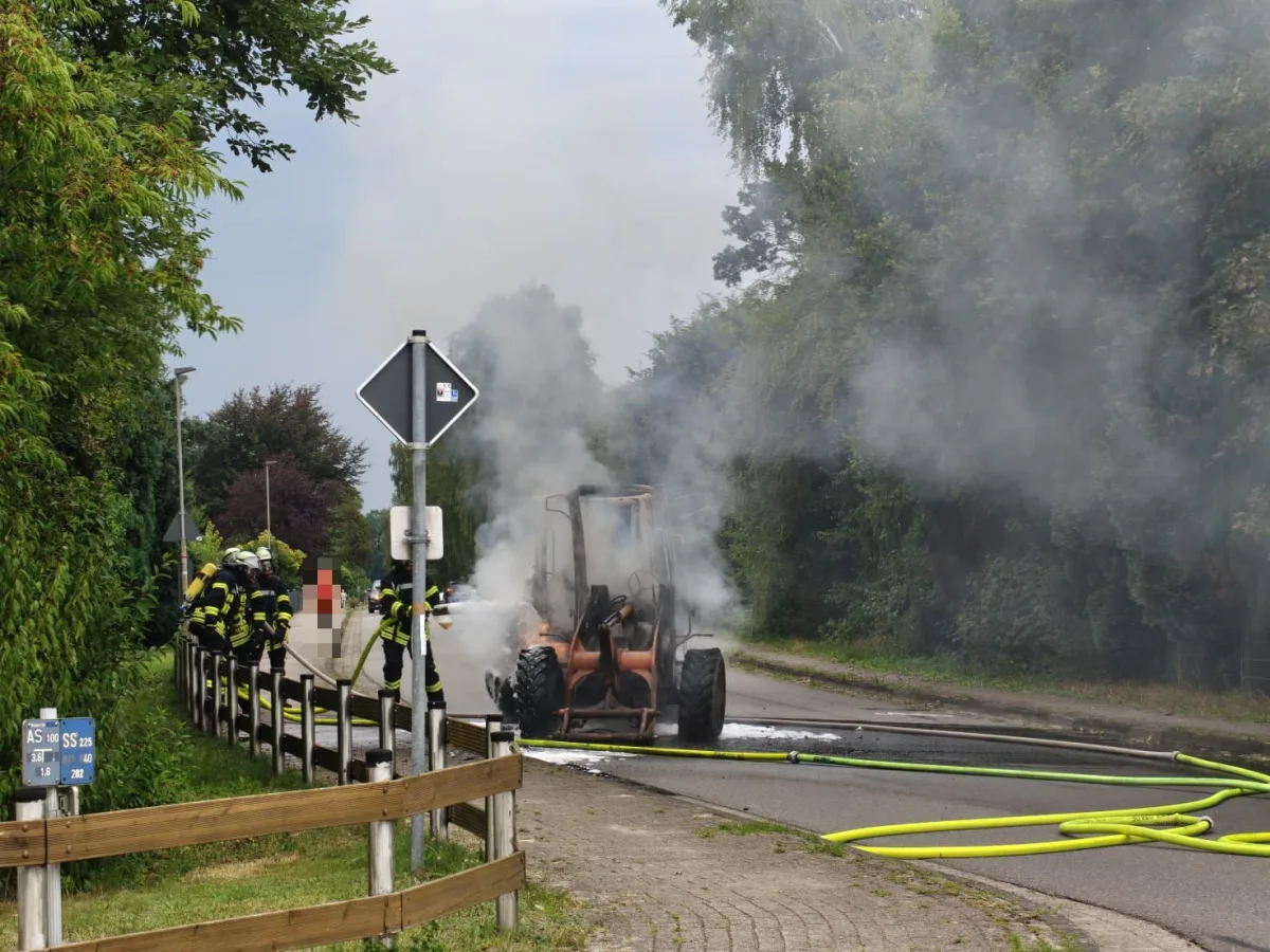 Ein brennender Bagger am Straßenrand wird von Feuerwehrleuten mit Schläuchen gelöscht. Rauch steigt auf, die Umgebung ist von Bäumen gesäumt.
