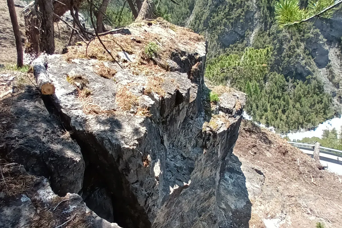 Ein steiler Felsvorsprung mit einer tiefen Kluft unterhalb, umgeben von Wald und mit einem Geländer im Hintergrund.
