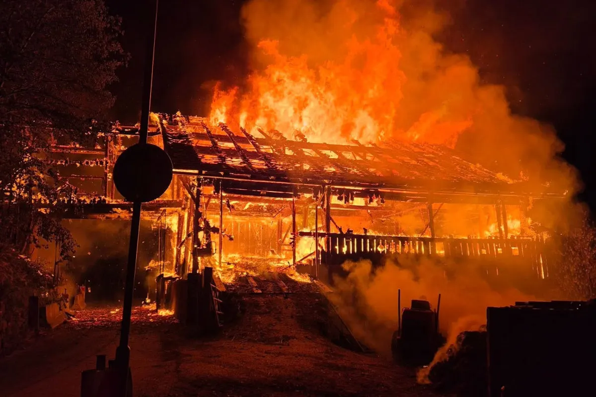 Ein großer Schuppen steht bei Nacht vollständig in Flammen, das Dach ist bereits eingestürzt. Der Vordergrund ist mit Rauch bedeckt.