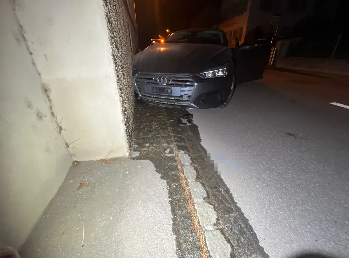 Ein dunkler Audi ist frontal gegen eine grob strukturierte Hauswand geprallt. Der Vorfall ereignete sich nachts auf einer gepflasterten Straße.
