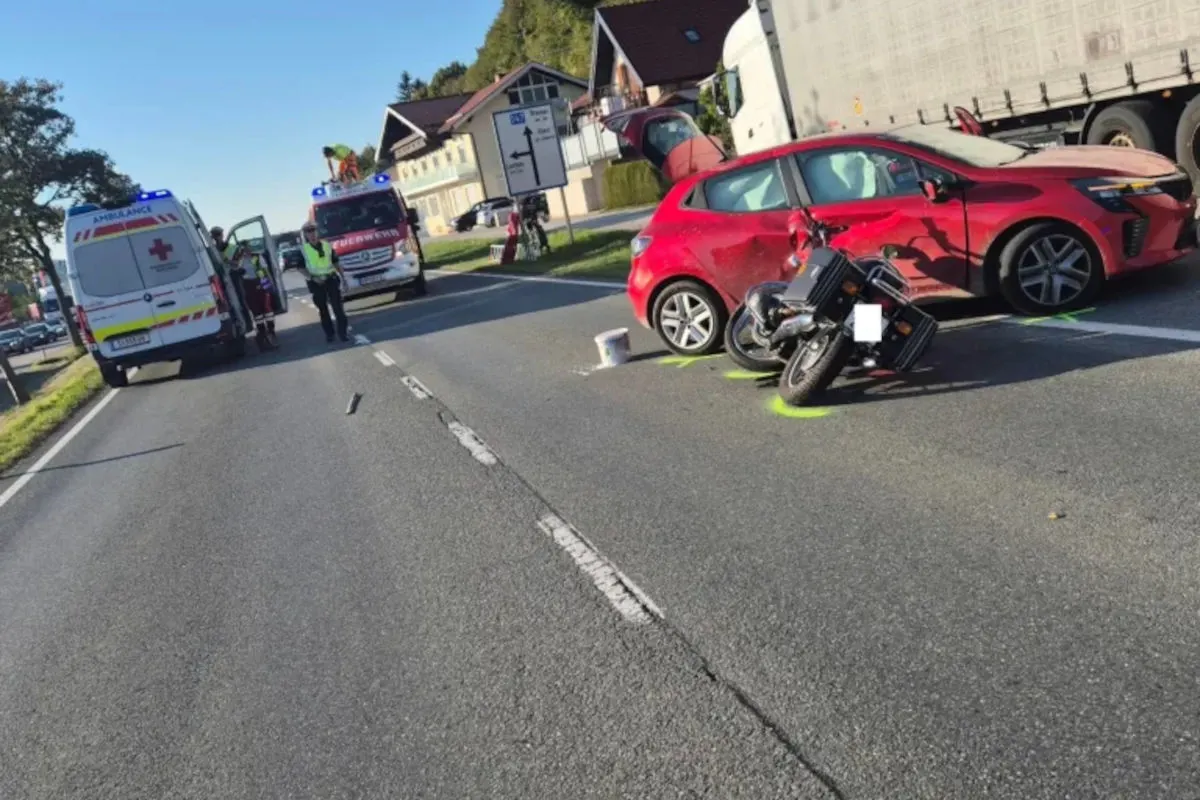 Ein beschädigtes rotes Auto und ein umgestürztes Motorrad stehen auf einer Straße, mit Rettungsfahrzeugen im Hintergrund.