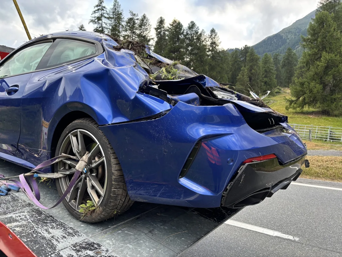 Ein stark beschädigter blauer Sportwagen steht auf einem Abschleppwagen. Die umgebende Landschaft zeigt Bäume und Hügel.