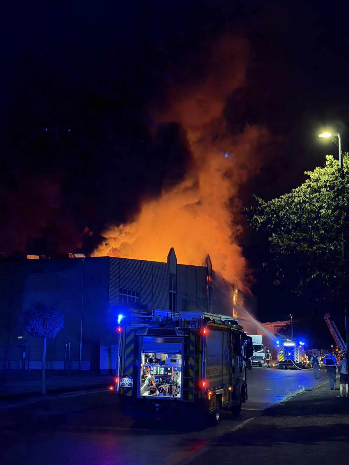 Mehrere Feuerwehrfahrzeuge sind bei Nacht im Einsatz vor einem großen brennenden Gebäude. Flammen und Rauch steigen in den Nachthimmel.