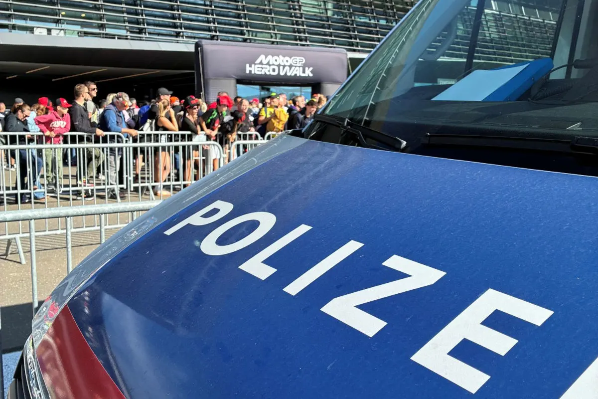 Ein Polizeifahrzeug steht im Vordergrund, während dahinter eine Menschenmenge bei einem Event sichtbar ist. Die Sonne scheint und es sind Sicherheitsabsperrungen vorhanden.