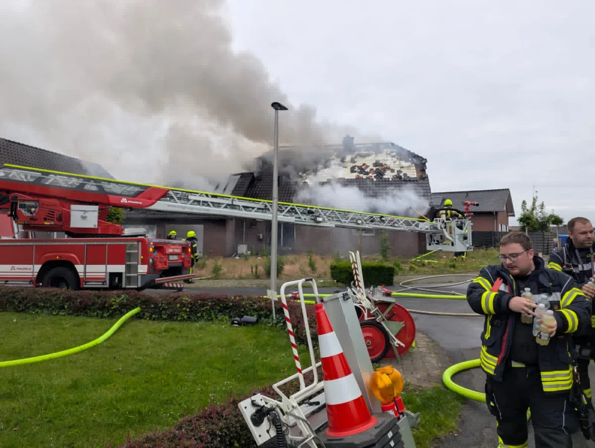Ein Feuerwehrmann ist auf einer Drehleiter, die auf das Dach eines Hauses gerichtet ist. Dichter Rauch steigt auf und weitere Feuerwehrleute sind vor Ort.