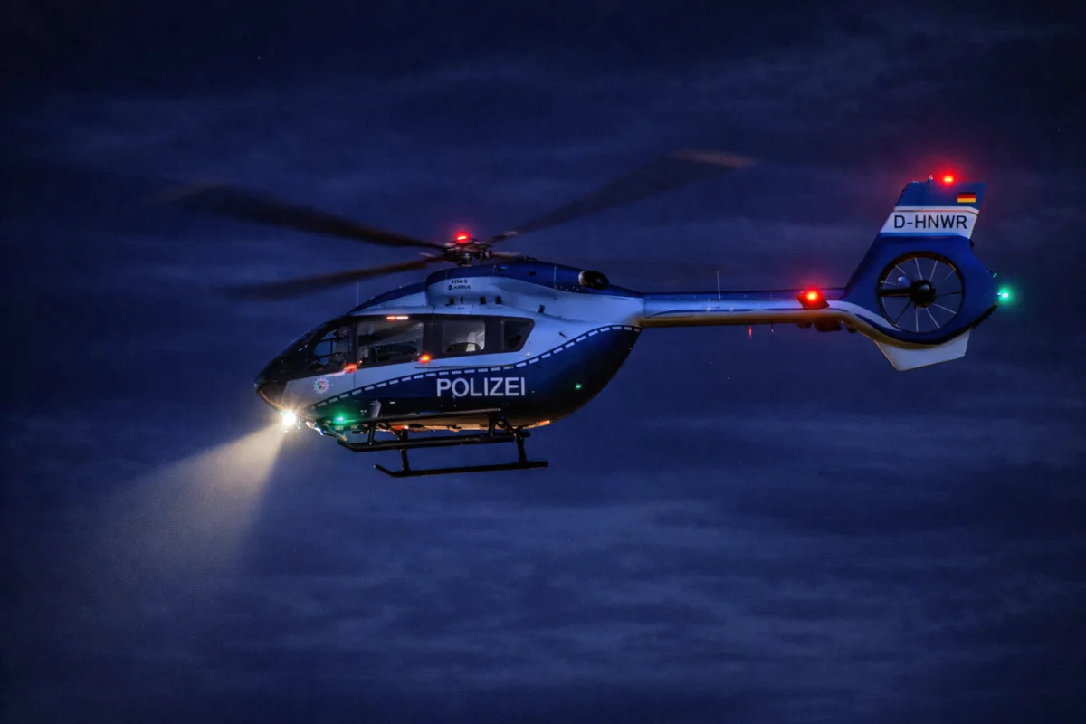 Ein Polizeihubschrauber mit eingeschaltetem Suchscheinwerfer fliegt in der Nacht am dunklen Himmel. Der Hubschrauber trägt die Aufschrift 'Polizei' und ist mit Lichtern ausgestattet.