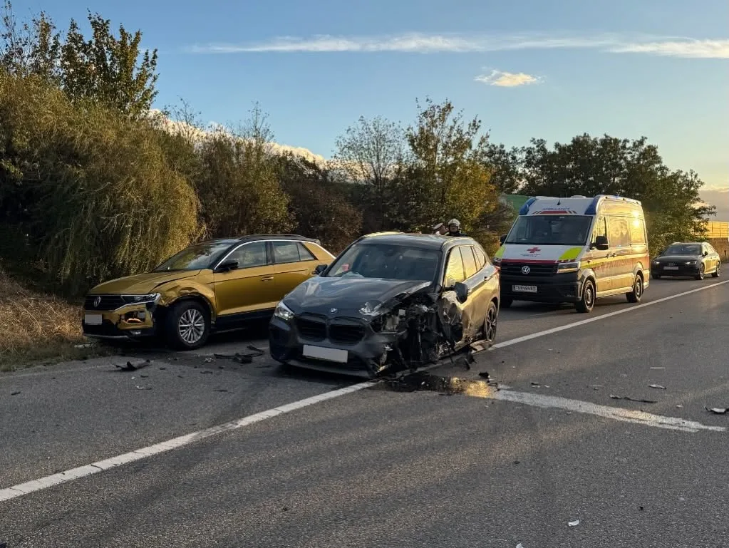 Ein schwarzes Auto mit erheblichen Frontschäden steht auf einer Landstraße. Daneben ein beschädigter, goldener SUV. Ein Rettungswagen ist auf der Straße sichtbar.