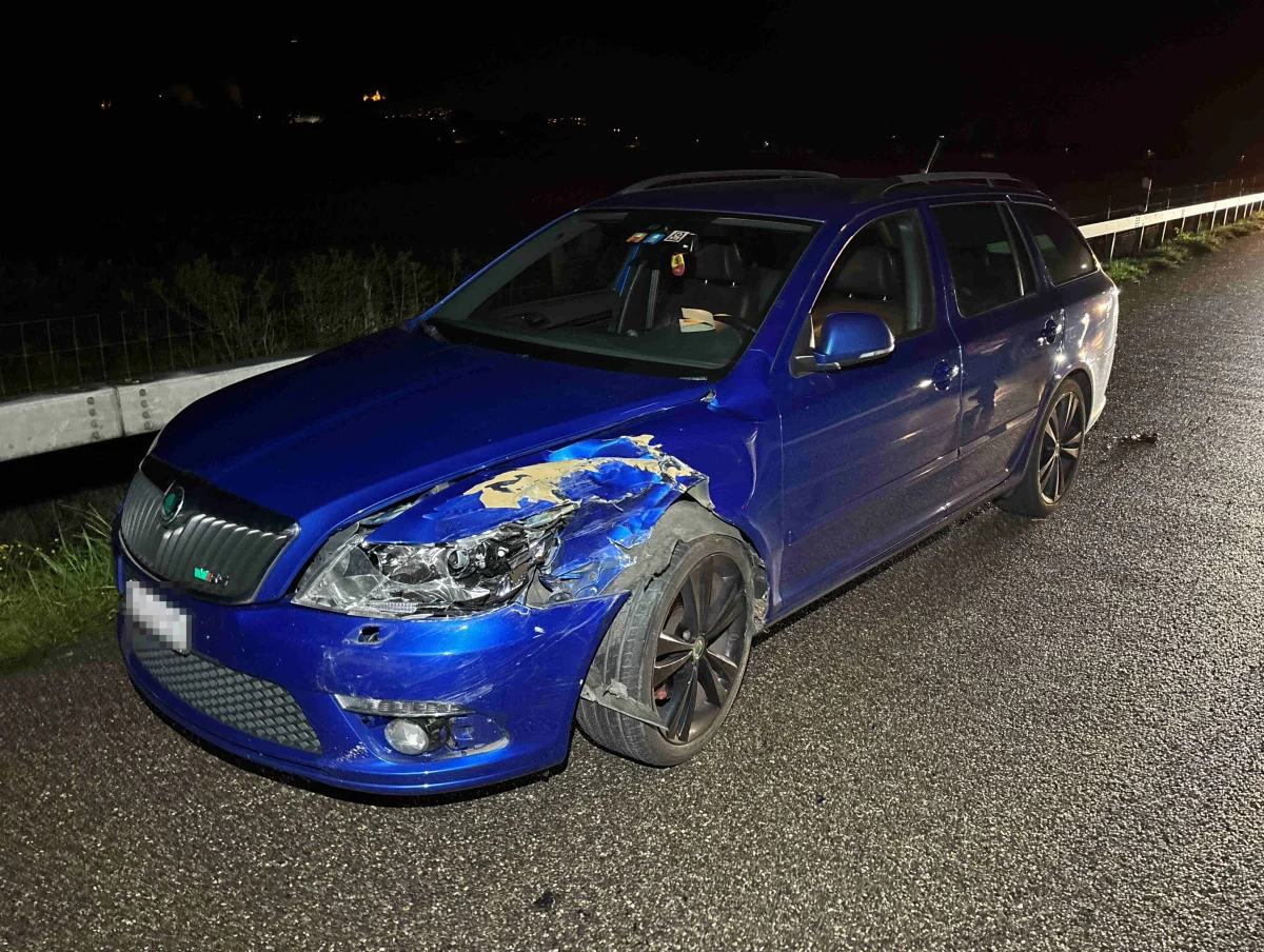 Ein blauer Kombi mit deutlichen Schäden an der vorderen Stoßstange steht bei Nacht am Straßenrand, beleuchtet durch Straßenlaternen.