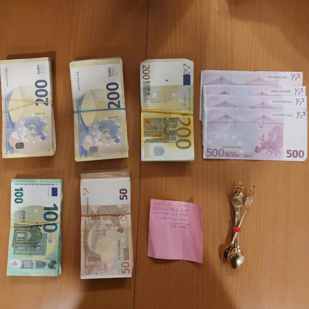 Auf einem Holztisch liegen mehrere Bündel Eurobanknoten unterschiedlicher Stückelungen sowie ein pinker Notizzettel und Schmuck.