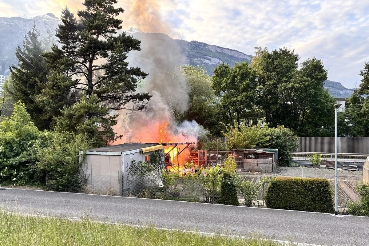 Ein Gartenhaus steht in Flammen, die Rauchwolken steigen in den Himmel. Im Hintergrund sind Bäume und Berge sichtbar.