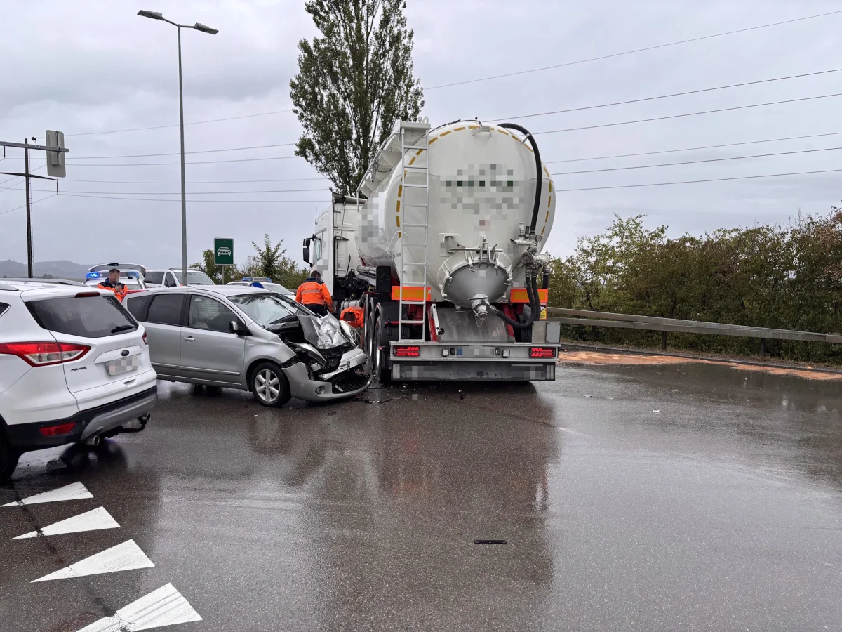 Ein silbernes Auto ist auf einer Straße bei nassem Wetter frontal mit einem weißen Tanklastwagen kollidiert. Mehrere Personen in orangenen Westen stehen um die Unfallstelle herum.