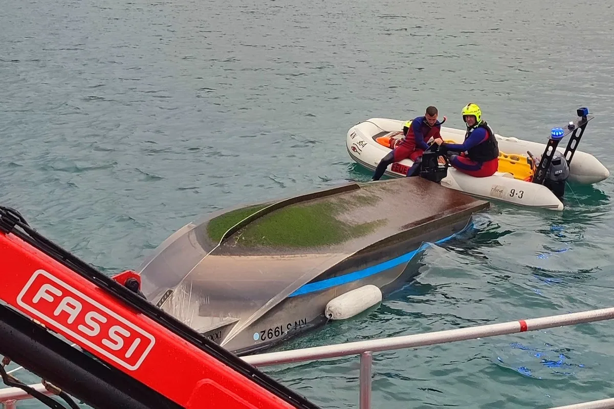 Zwei Rettungskräfte in einem Schlauchboot arbeiten am gekenterten Boot auf einem Gewässer. Ein Kran ist im Vordergrund sichtbar.