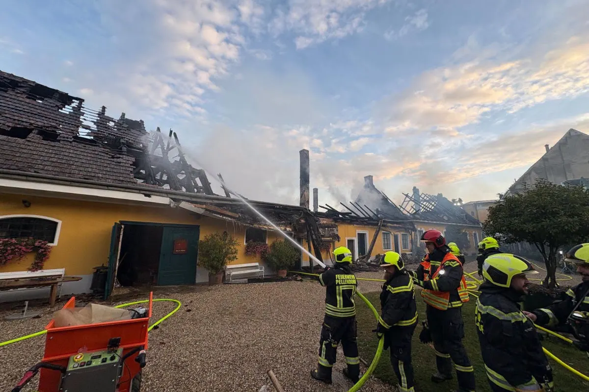 Mehrere Feuerwehrleute in Schutzanzügen löschen mit Schläuchen ein Gebäude, dessen Dach durch ein Feuer zerstört wurde. Rauch steigt vom Dach auf, während der Himmel teilweise bewölkt ist.