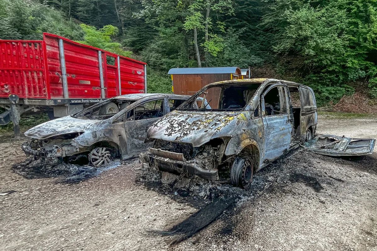 Zwei stark beschädigte Fahrzeuge stehen nebeneinander auf einer Waldstraße. Sie sind ausgebrannt und von Ruß bedeckt.