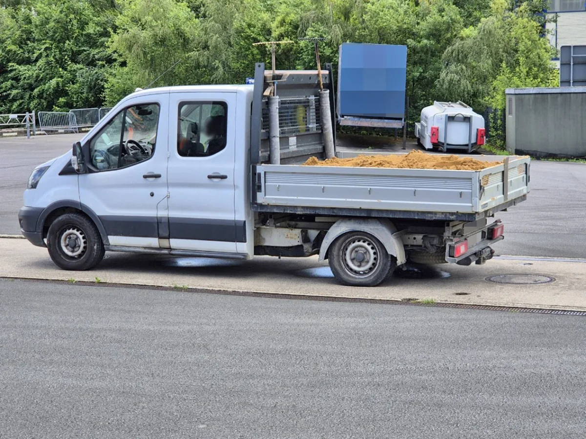 Ein weißer Transporter steht seitlich geparkt und ist mit Sand auf der offenen Ladefläche beladen, umgeben von Grünflächen.