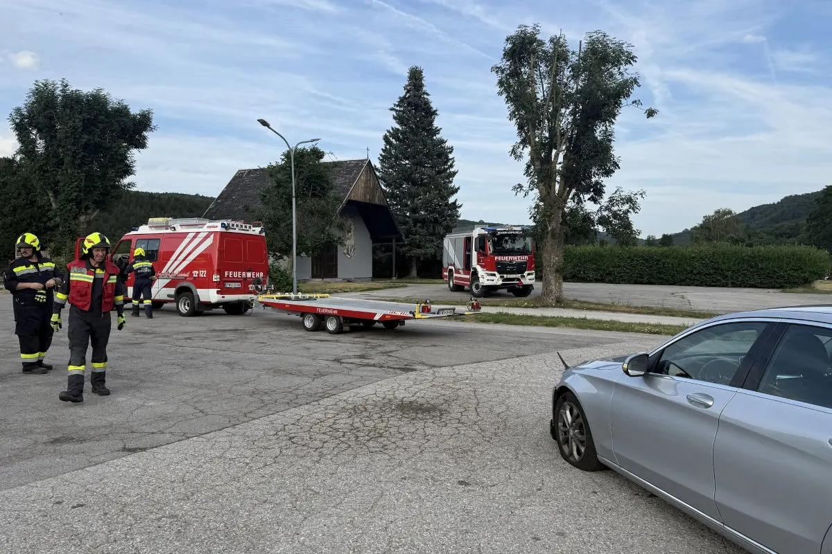 Mehrere Feuerwehrleute stehen in Einsatzkleidung neben zwei Feuerwehrfahrzeugen und einem Anhänger auf einem Parkplatz.