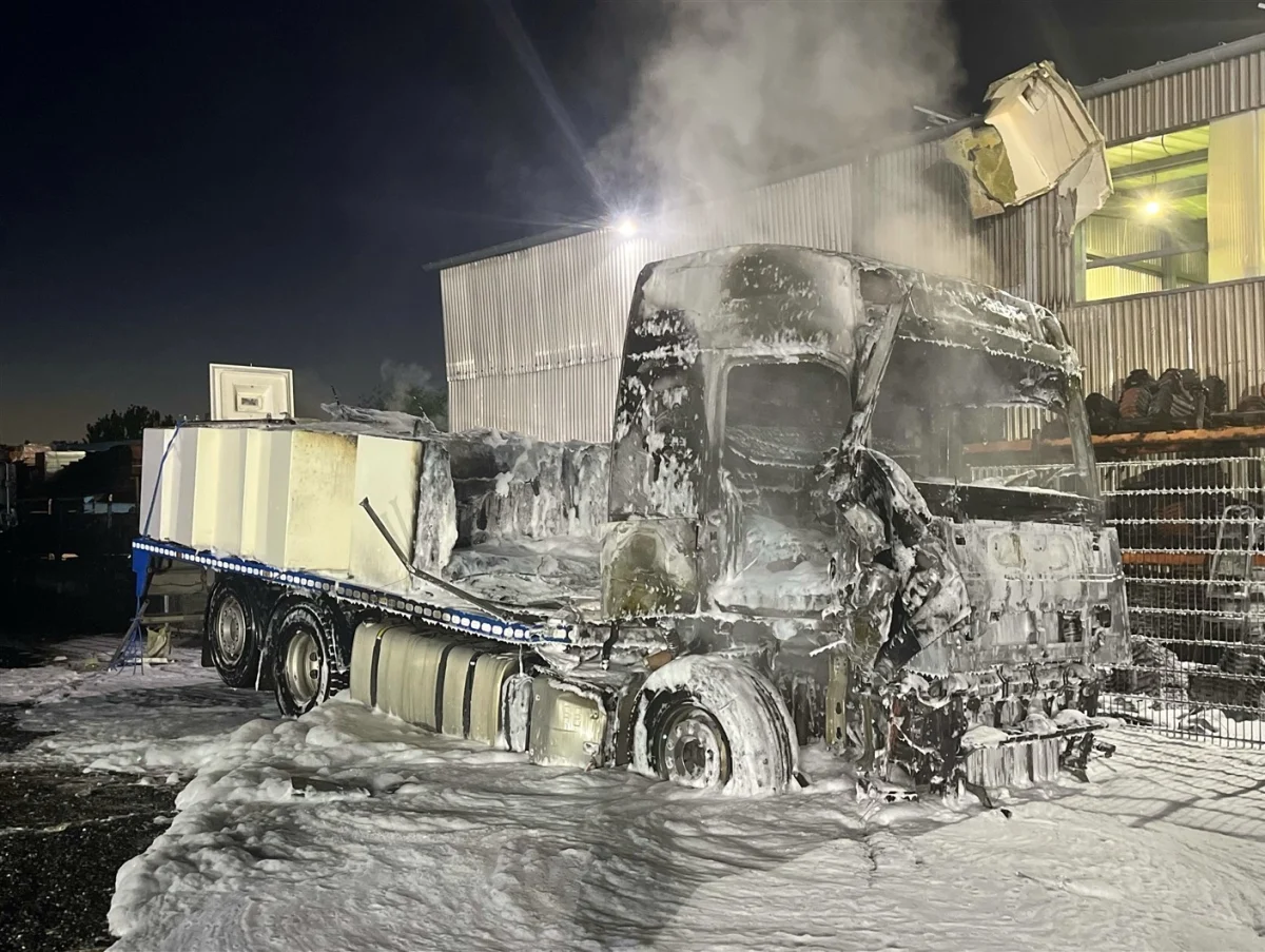 Ein LKW steht verbrannt und mit Löschschaum bedeckt in einem Industriegebiet. Es ist Nacht, und im Hintergrund ist ein beleuchtetes Gebäude sichtbar.