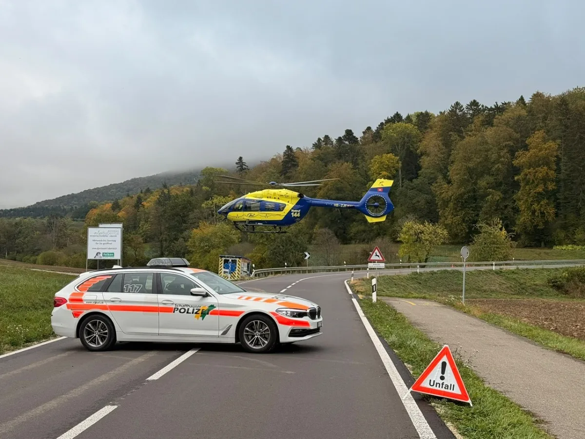 Ein weißes Polizeifahrzeug blockiert eine Landstraße, während ein gelb-blauer Rettungshubschrauber in der Nähe landet. Ein Unfall-Warnschild ist sichtbar.