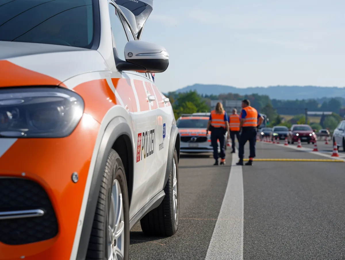 Ein weißes und orangefarbenes Polizeifahrzeug steht auf einer Autobahn. Beamte in orangefarbenen Westen stehen in der Ferne auf der Straße. Die Straße ist mit Kegeln abgesperrt.