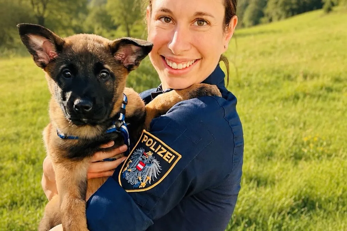 Eine Frau in Polizeiuniform hält einen Welpen in ihren Armen, und sie stehen auf einer grünen Wiese unter freiem Himmel.