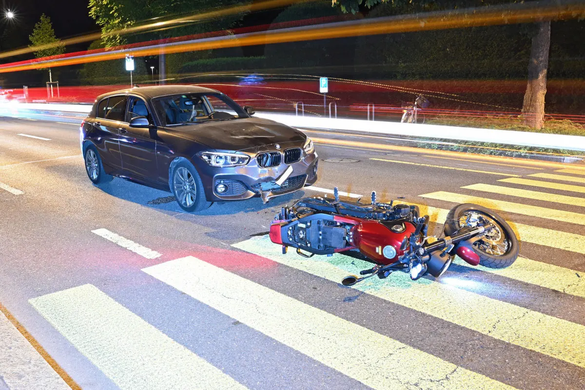 Ein schwarzer BMW steht nahe einem umgefallenen roten Motorrad auf einer beleuchteten Straße bei Nacht. Im Hintergrund sind Lichtstreifen vorbeifahrender Fahrzeuge sichtbar.
