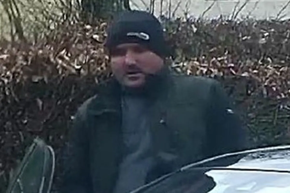 Eine Person in dunkler Kleidung und Mütze steht neben einem Auto vor einem kahlen Gebüsch. Es scheint kühl zu sein.