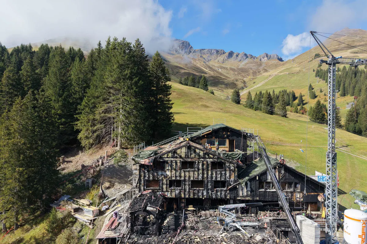 Ein abgebranntes, mehrstöckiges Gebäude in einer alpinen Umgebung mit grünen Wiesen und Bergen im Hintergrund. Arbeiter und ein Kran sind vor Ort sichtbar.
