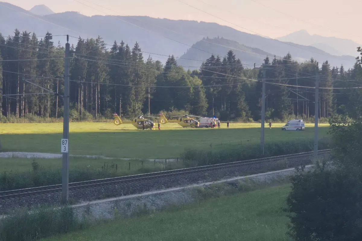 Zwei gelbe Rettungshubschrauber stehen auf einer Wiese, begleitet von einem Rettungsfahrzeug. Im Hintergrund sind Bäume und Berge zu sehen.