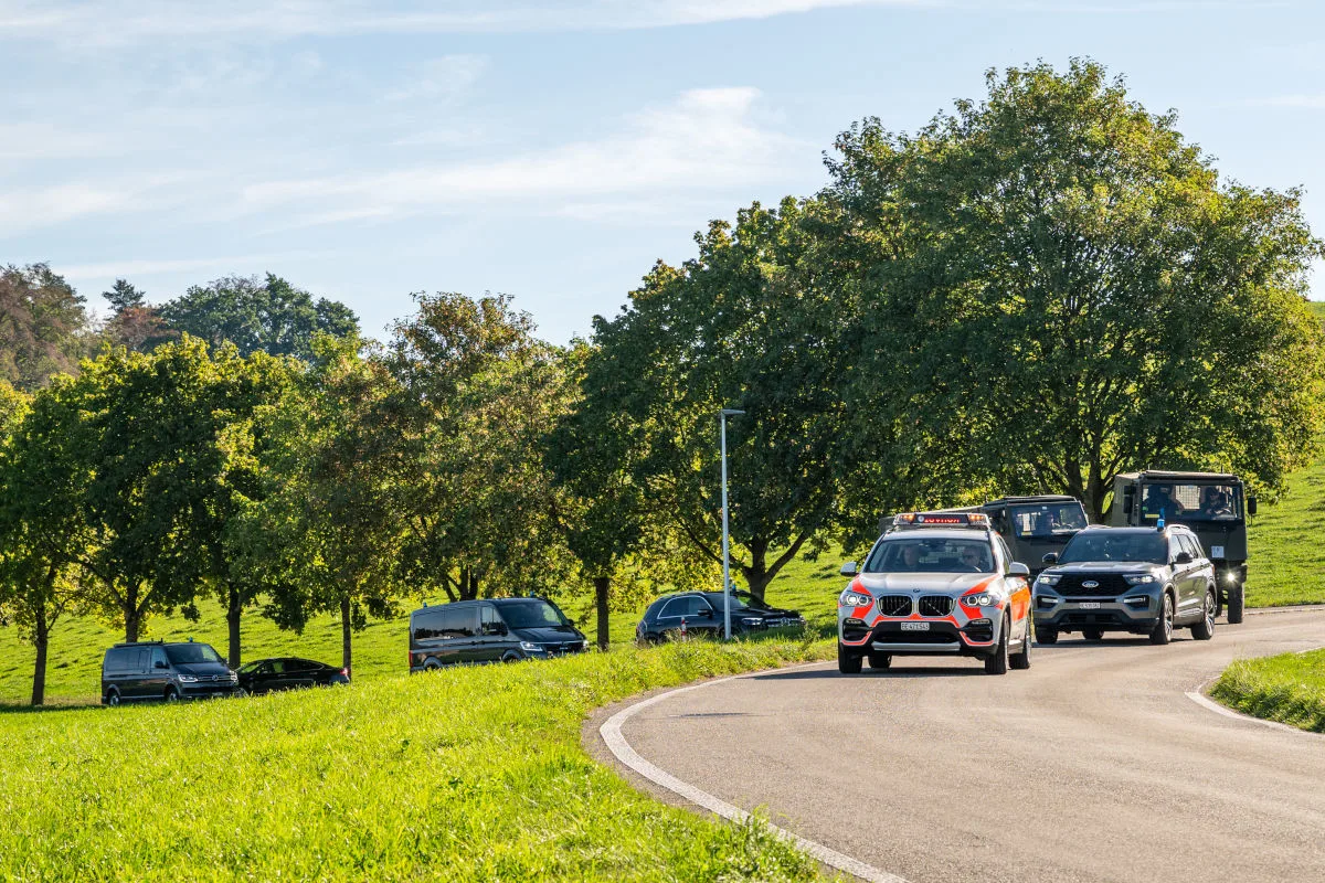 Ein Rettungswagen fährt auf einer kurvigen Landstraße, gefolgt von mehreren Fahrzeugen. Im Hintergrund sind grüne Bäume und ein klarer Himmel zu sehen.