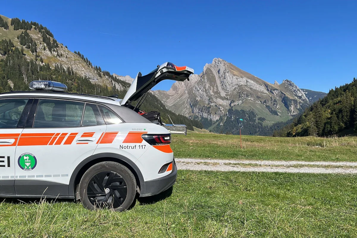 Ein Polizeifahrzeug mit geöffneter Heckklappe steht auf einer Wiese vor einer beeindruckenden Bergkulisse unter blauem Himmel.