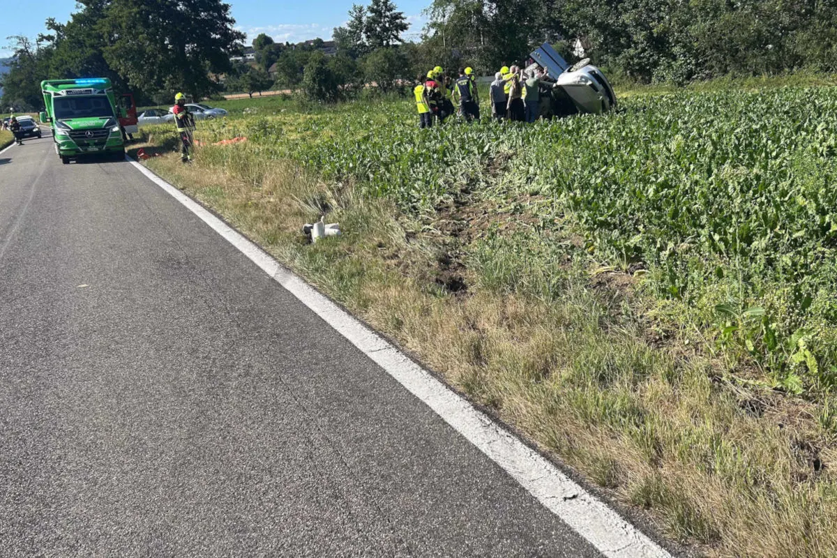 Eine Gruppe von Einsatzkräften in gelben Helmen steht bei einem umgestürzten Fahrzeug im Feld. Ein Rettungsfahrzeug ist auf der Straße.