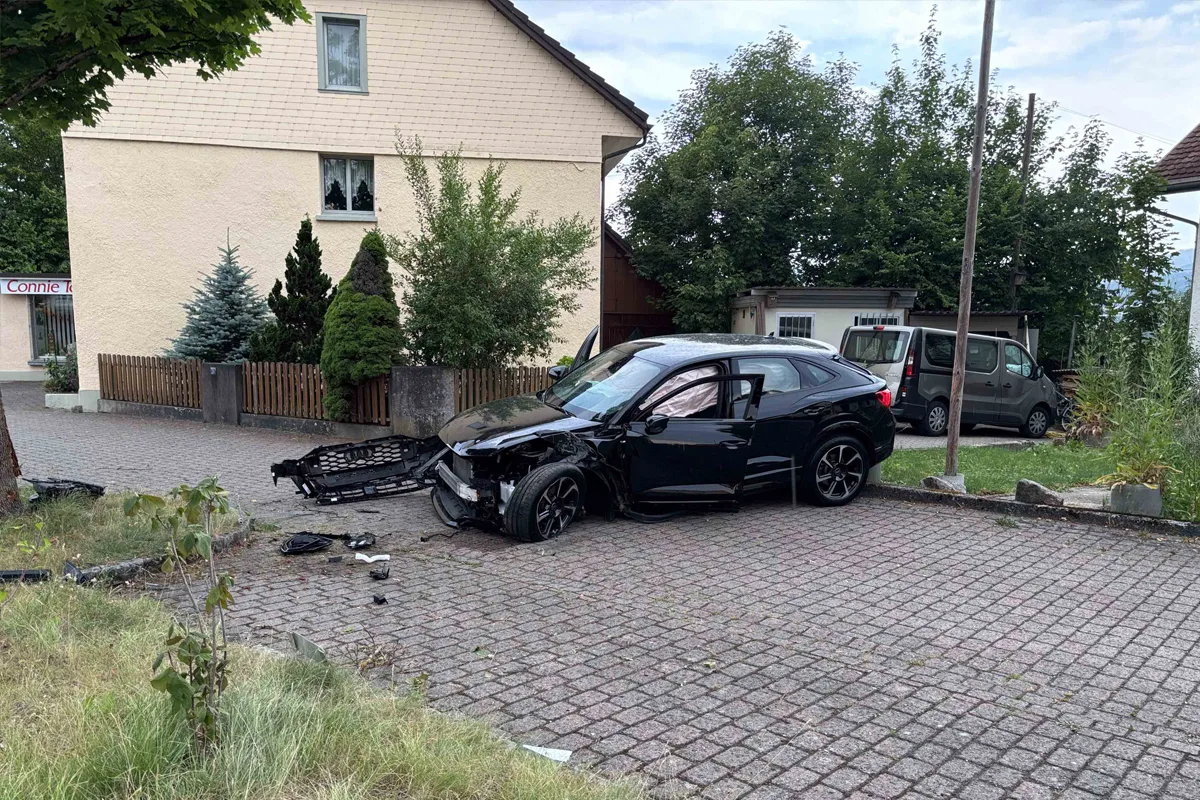 Ein schwarzes Auto steht mit erheblichem Frontschaden auf einem gepflasterten Gehweg vor einem Haus. Auch Fahrzeugteile sind auf dem Boden verteilt.