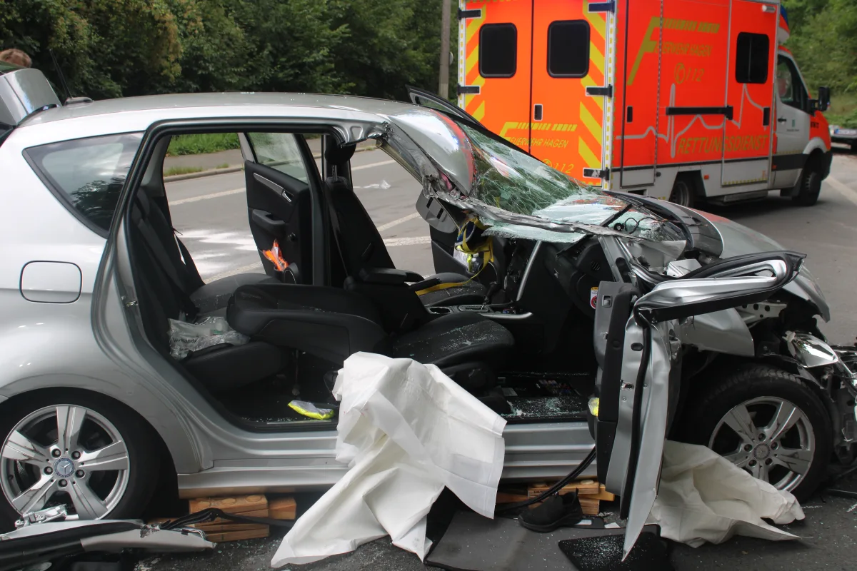 Ein stark beschädigtes silbernes Auto steht am Straßenrand mit geöffneten Türen. Im Hintergrund ist ein Rettungswagen sichtbar.