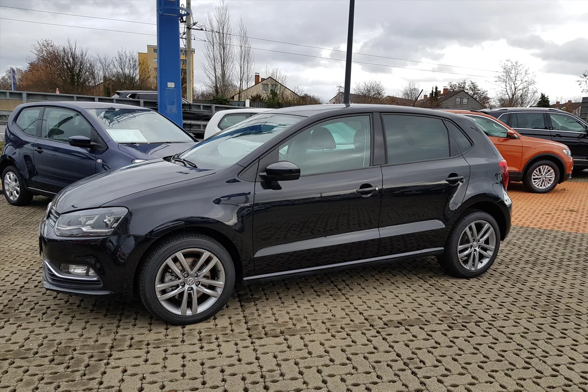 Auf dem Bild ist ein schwarzer Volkswagen auf einem gepflasterten Parkplatz zu sehen, umgeben von anderen Autos.