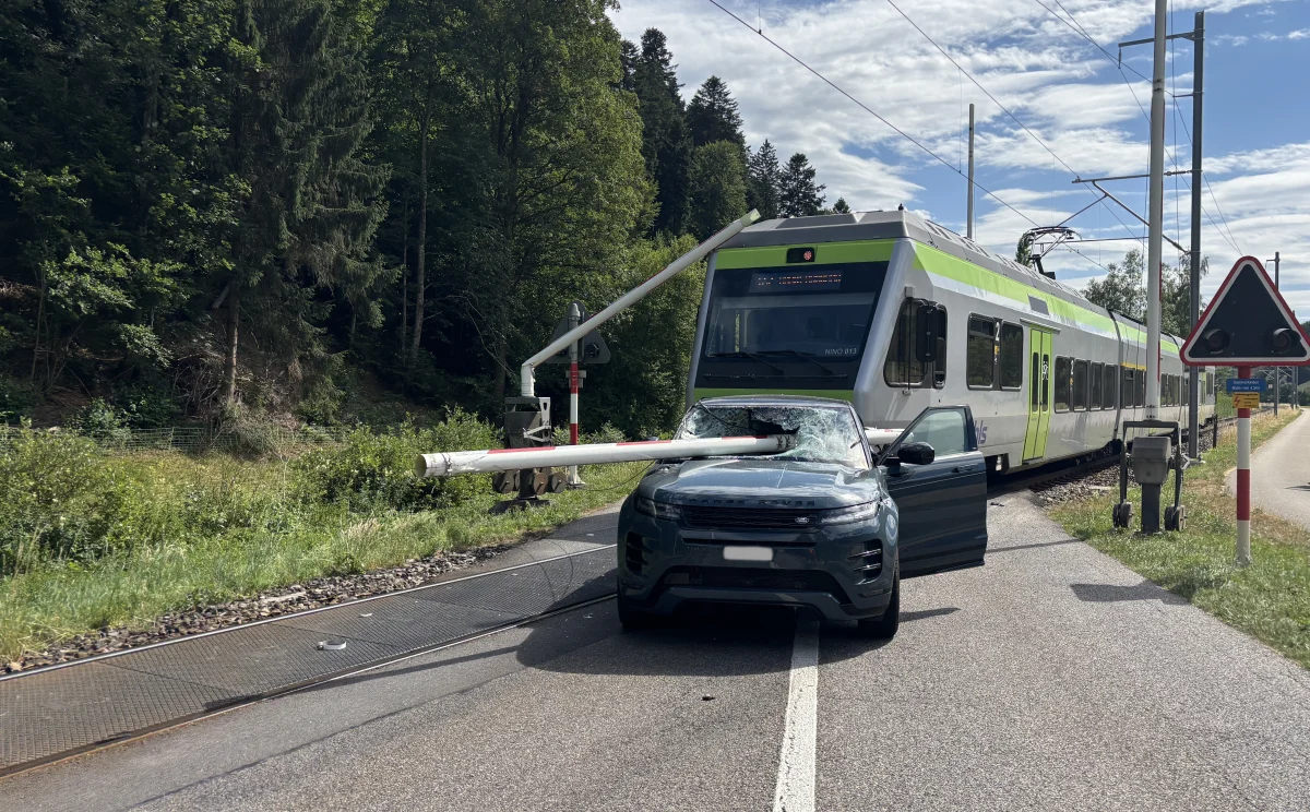 Ein SUV kollidierte mit einer Schranke am Bahnübergang, während ein Zug im Hintergrund sichtbar ist. Das Auto scheint beschädigt, die Schranke durch die Windschutzscheibe gedrungen.
