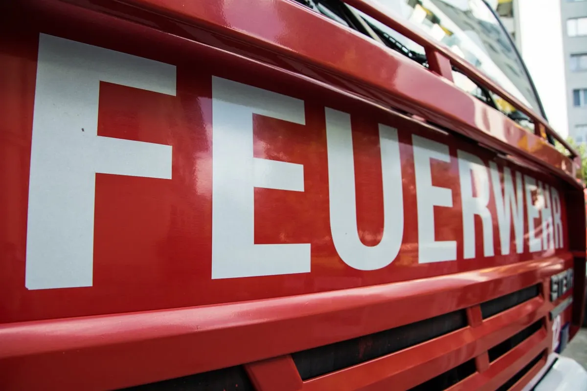 Das Bild zeigt eine Nahaufnahme der Vorderseite eines roten Feuerwehrautos mit dem großen weißen Schriftzug 'FEUERWEHR'. Rechts im Hintergrund sind unscharfe Gebäude zu sehen.