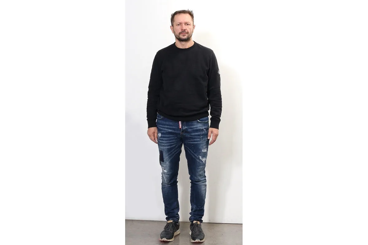 Ein Mann steht vor einer weißen Wand. Er trägt einen dunkelblauen Pullover und blaue Jeans.