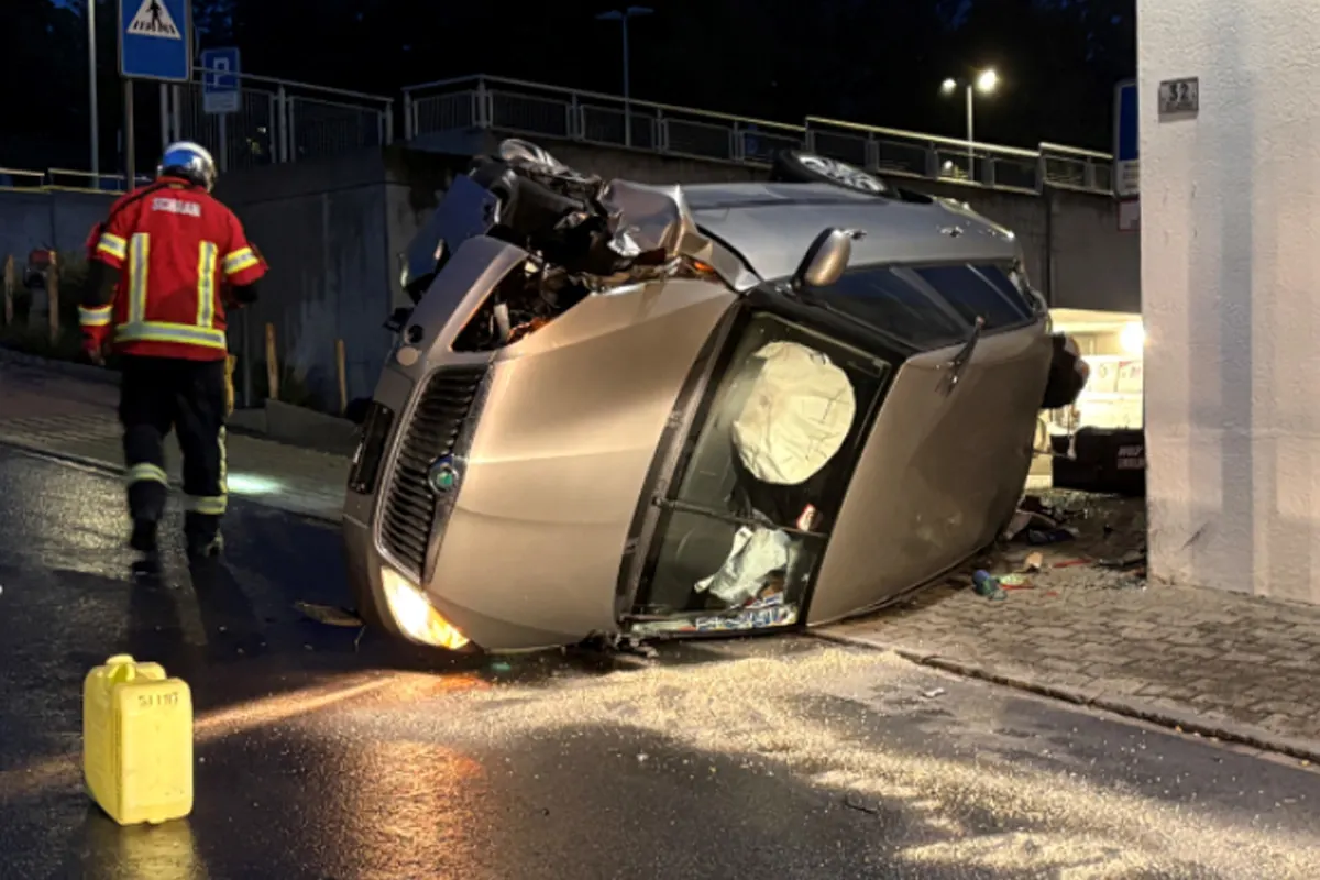 Ein Auto liegt seitlich auf einer nassen Straße mit einem aktivierten Airbag. Ein Feuerwehrmann in roter Uniform steht daneben.