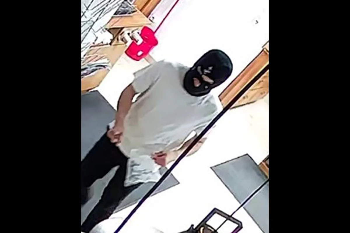 Eine Person mit schwarzer Maske und weißem T-Shirt steht in einem Geschäft. Im Hintergrund sind Regale sichtbar.