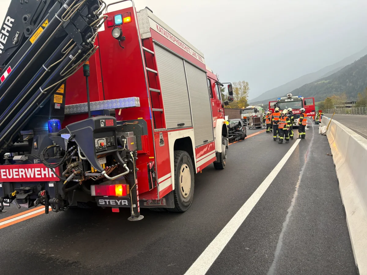 Zwei rote Feuerwehrfahrzeuge stehen auf einer Straße, umgeben von Feuerwehrleuten in orangefarbenen Westen. Ein demoliertes Auto ist im Hintergrund sichtbar.