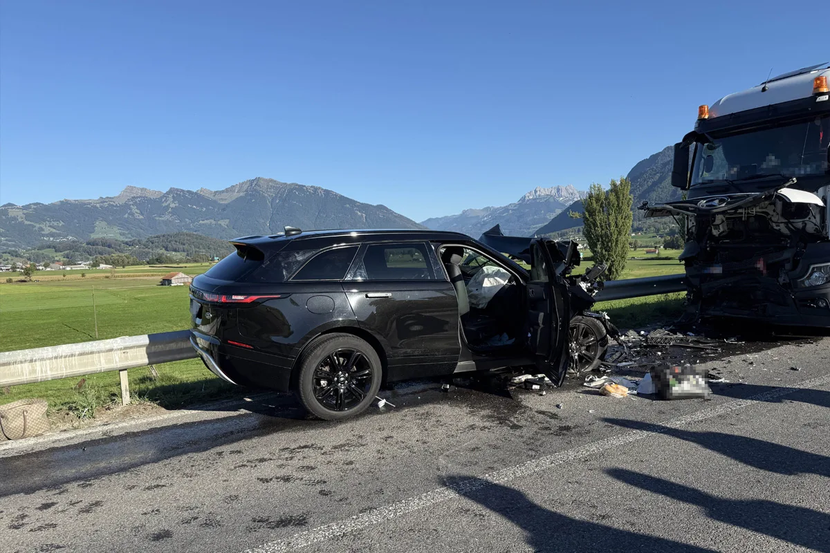 Ein schwarzer SUV steht beschädigt nach einem Frontalzusammenstoß mit einem Lastwagen auf einer Landstraße. Die Landschaft im Hintergrund zeigt eine Bergkulisse.