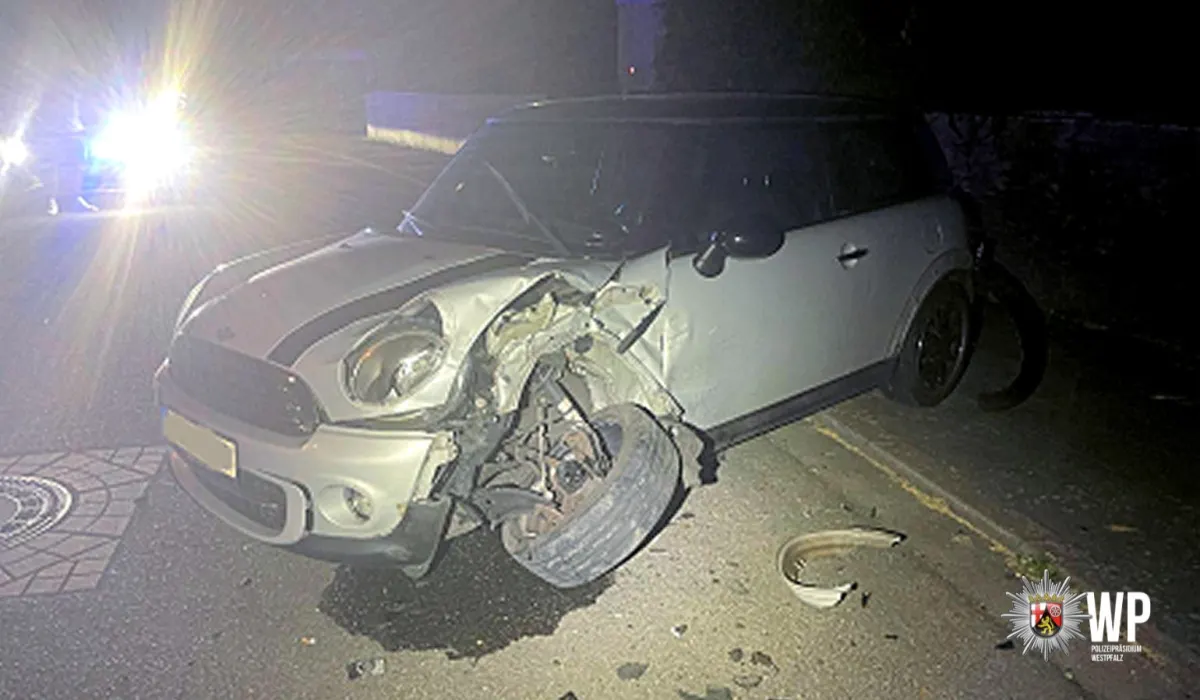 Ein silberner Mini Cooper zeigt schwere Frontschäden, das linke Vorderrad ist stark verbogen. Es ist nacht, und ein Polizeifahrzeug mit Blaulicht ist im Hintergrund zu sehen.